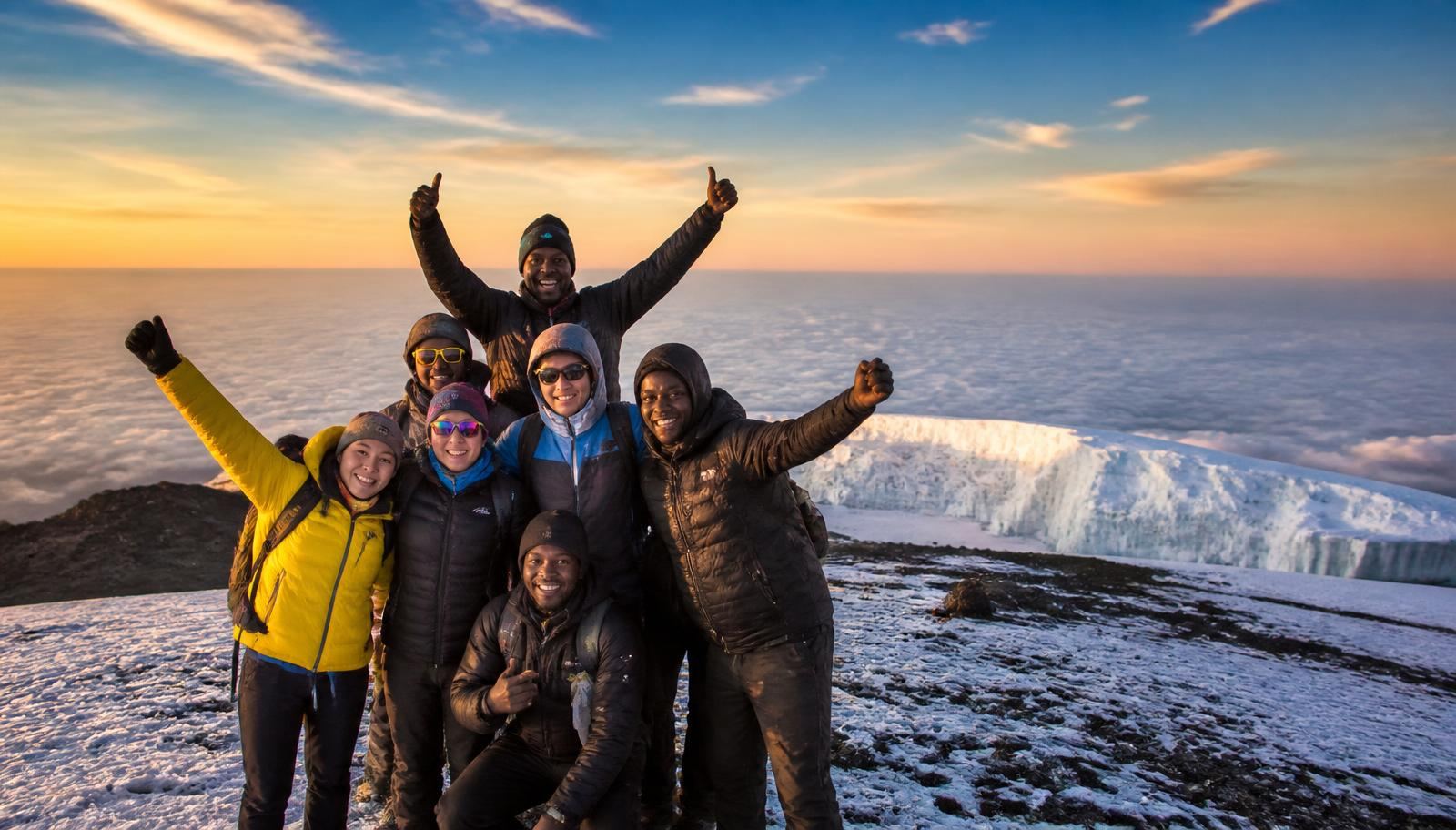 International climbers celebrating with Tanzanian mountain guides at Uhuru Peak on the snow-covered summit of Mount Kilimanjaro at sunrise above the clouds