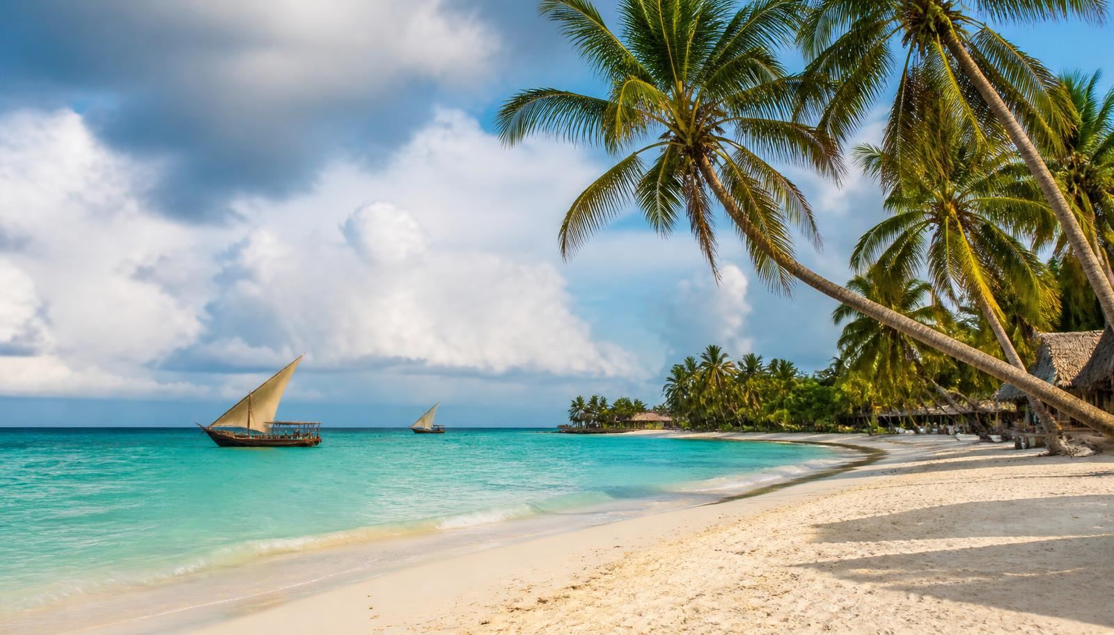Panoramic Zanzibar beach with white sand, turquoise Indian Ocean water, leaning palm trees and traditional wooden dhow boats sailing under bright tropical sunshine and seasonal clouds